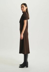 Robe midi marron en tissu lisse avec des manches bouffantes et une ceinture à la taille, associée à des bottes noires montantes et des accents en cuir noir élégants.