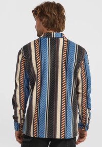 Hombre con cabello castaño ondulado de espaldas, vistiendo una camisa de manga larga con rayas verticales en azul, marrón, negro, crema y blanco.