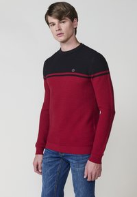 Maglione lavorato a maglia rosso e nero con scollatura a girocollo, caratterizzato da righe orizzontali e un piccolo logo sul petto. Maniche lunghe e polsini a coste.