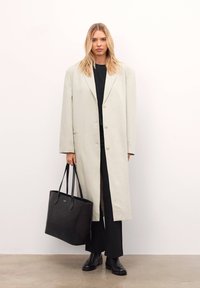 Világosszürke hosszú kabát nagy gombokkal, fekete felső és nadrág fölött viselve, fekete bevásárlótáskával és fekete cipővel, semleges háttér előtt.