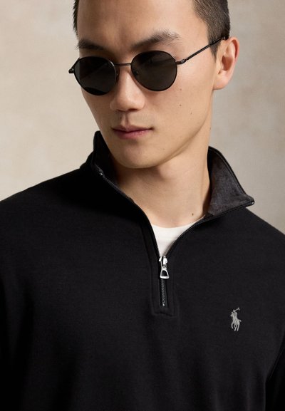 Joven con gafas de sol negras redondas y un suéter negro con cremallera hasta el pecho y un pequeño logo bordado en el pecho.