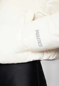 Svijetlosmeđi puffer jakna s visokim ovratnikom i rebrastim manžetama. Ima suptilan sivi "Fabletics" logo na rukavu. Glatka tekstura.
