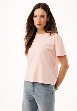 Mujer con cabello largo y castaño, vistiendo una camiseta de manga corta de color rosa claro y vaqueros azul claro, de pie frente a un fondo blanco.