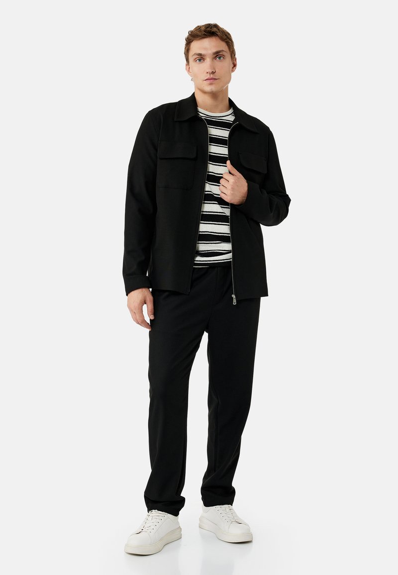 Joven con chaqueta negra sobre una camiseta de rayas blancas y negras, pantalones negros y zapatillas blancas, de pie frente a un fondo liso.