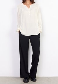 Blouse beige clair avec un col henley à boutons et des manches longues, associée à un pantalon large noir et des chaussures noires.