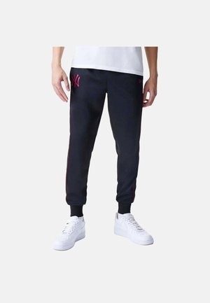 Personne portant un pantalon de jogging noir avec des bandes latérales roses et un logo sur la cuisse gauche, des baskets blanches et un haut blanc.