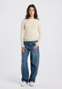 Woman standing wearing a long-sleeve cream lace top, loose blue jeans, and white sneakers against a plain light background.