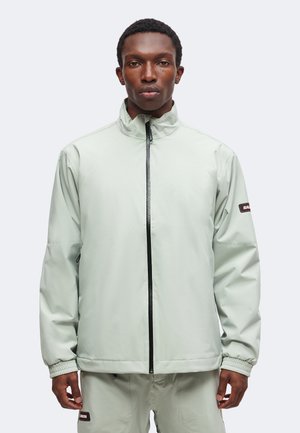 Homme portant une veste zippée gris clair à col montant et un pantalon assorti sur un fond uni.