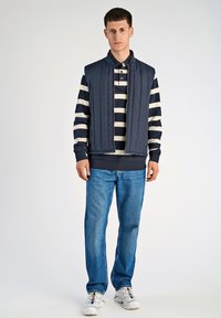 Marinkviltad väst över en randig långärmad skjorta, kombinerad med blå jeans och vita skor med röda och blå accenter. Enkelt, avslappnat design.