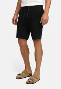 Hombre vestido con pantalones cortos negros de cordones y sandalias de doble tirante en color beige, de pie contra un fondo blanco liso.