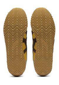 Hnědé a žluté podrážky tenisek s vlnitým vzorem, s logem na levé podrážce. Materiál vypadá gumově a má texturu.