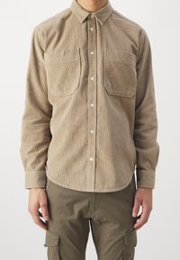Camicia in velluto a coste marrone con colletto button-down, due tasche sul petto e chiusura a bottoni. Maniche lunghe con dettagli ai polsini.