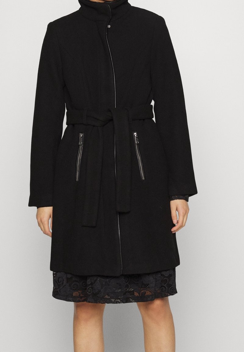 Cappotto in lana nera con alto colletto, maniche lunghe, chiusura frontale con zip, cintura in vita e due tasche con zip. Texture liscia.