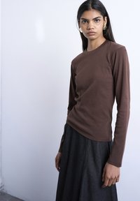 Γυναίκα με μακριά σκούρα μαλλιά που φοράει χρυσά σκουλαρίκια, καφέ μπλούζα με μακριά μανίκια και σκουρόχρωμη φούστα με ψηλή μέση, στέκεται μπροστά σε ένα απλό ανοιχτό φόντο.