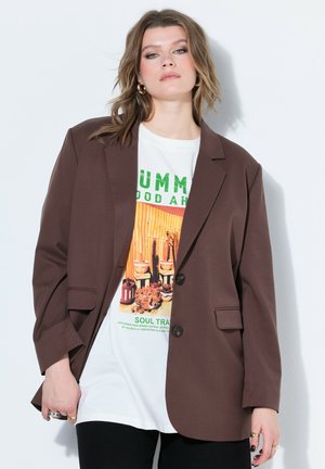 Młoda kobieta ubrana w brązową marynarkę na białej koszulce z nadrukiem, stojąca na tle jasnego, gładkiego tła z neutralnym wyrazem twarzy.