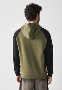 Hoodie vert olive avec des manches noires, doté d'une capuche à cordon de serrage et de poignets côtelés. Tissu doux et texturé avec une coupe décontractée.
