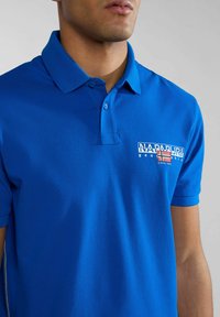 Camisa polo azul hecha de tejido texturizado, con un cuello, dos botones y un logo en el lado izquierdo. Diseño simple y mangas cortas.