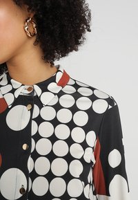 Černá halenka s velkým vzorem bílých a červených puntíků, s límečkem a zlatými knoflíky. Měkká, hladká textura materiálu.