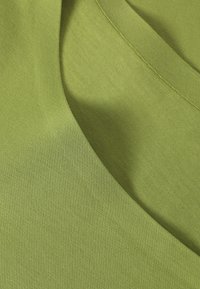 Zelená tkanina s hladkým texturem, s kulatým výstřihem a subtilními detaily šití. Materiál se zdá lehký a měkký.