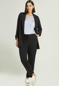 Blazer negro a medida sobre una blusa de satén plateado claro, combinado con pantalones jogger negros y zapatillas metálicas plateadas. Diseño minimalista, look casual.