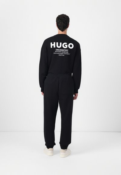 Sweatshirt noir et pantalon assorti avec taille élastique, présentant le logo blanc "HUGO" au dos et une poche arrière. Tissu lisse, coupe décontractée.