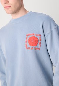 Light blue sweatshirt featuring a textured red circular graphic and bold white text. Round neckline and ribbed cuffs. Casual fit.