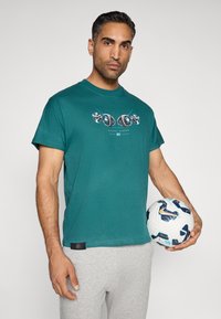 Camiseta de algodão verde-água com um design gráfico no peito. Modelo segura uma bola de futebol multicolorida. Calças cinzas visíveis.
