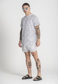 Camisa blanca de manga corta y pantalones cortos con un patrón repetido de logotipos negros, combinados con sandalias negras y gafas de sol, mostrando tatuajes en las piernas.