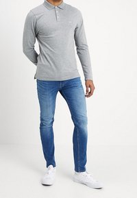 Man i en ljusgrå långärmad polo, blå slim-fit jeans och vita sneakers, står med en hand bakom ryggen.