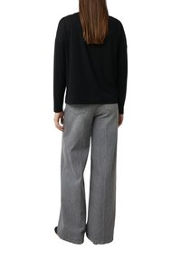 Frau steht mit dem Rücken zur Kamera, trägt ein schwarzes Langarmshirt und hoch taillierte weite graue Jeans vor weißem Hintergrund.