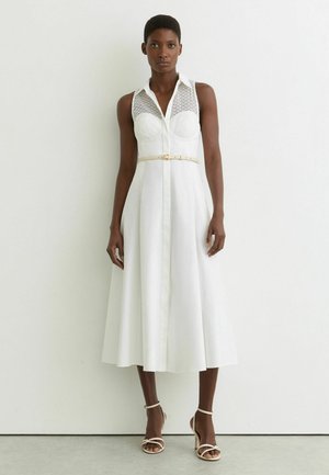 Kobieta ubrana w białą, bezrękawnikową sukienkę midi z koronkowym gorsetem, beżowym cienkim paskiem oraz beżowe sandały na wysokim obcasie ze rzemykami, stojąca na tle białej ściany.