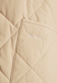 Beiger, gestepptes Material mit Rautenmotiv; verfügt über eine Vordertasche und ein gesticktes "Barbour"-Logo in weißem Faden. Glatte Textur.