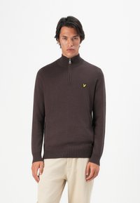 Mann iført en mørkebrune quarter-zip genser med en gul logo på brystet og lysebeige bukser, stående mot en hvit bakgrunn.