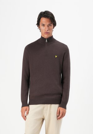 Homme portant un pull zippé marron foncé avec un logo jaune sur la poitrine et un pantalon beige clair, se tenant contre un fond blanc.