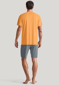 Oranje katoenen t-shirt gecombineerd met gestreepte blauwe shorts met geometrische ontwerpen. Gezicht vanaf de achterkant, model blootsvoets.