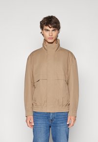 Chaqueta beige ligera con cuello alto, cierre de cremallera en la parte delantera y cinturilla elástica. Cuenta con un bolsillo horizontal en el pecho y un ajuste relajado.