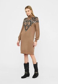 Brun tröjklänning med hög krage, intrikat svart broderi och långa ärmar. Bärs med svarta knähöga stövlar.