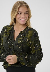 Junge Frau mit blonden Haaren lächelt, trägt eine dunkelgrüne und schwarze Blumenbluse mit langen Ärmeln, die Hände vor sich gefaltet vor einem schlichten grauen Hintergrund.