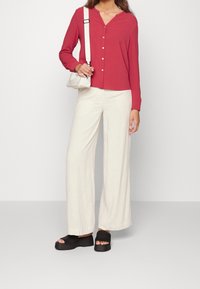 Camicetta rossa a maniche lunghe con bottoni, pantaloni larghi color crema e sandali con plateau neri. Include una piccola borsa a tracolla bianca.