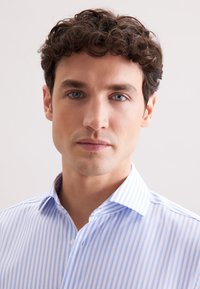 Lichtblauw en wit gestreept shirt met een klassieke kraag, voorzien van een knoopsluiting en een gladde textuur. Model toont de pasvorm van het shirt.