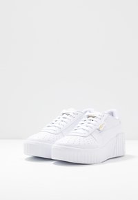 Baskets blanches à lacets avec une semelle plateforme texturée. Elles présentent une tige perforée, un col rembourré et un accent logo doré sur le côté.