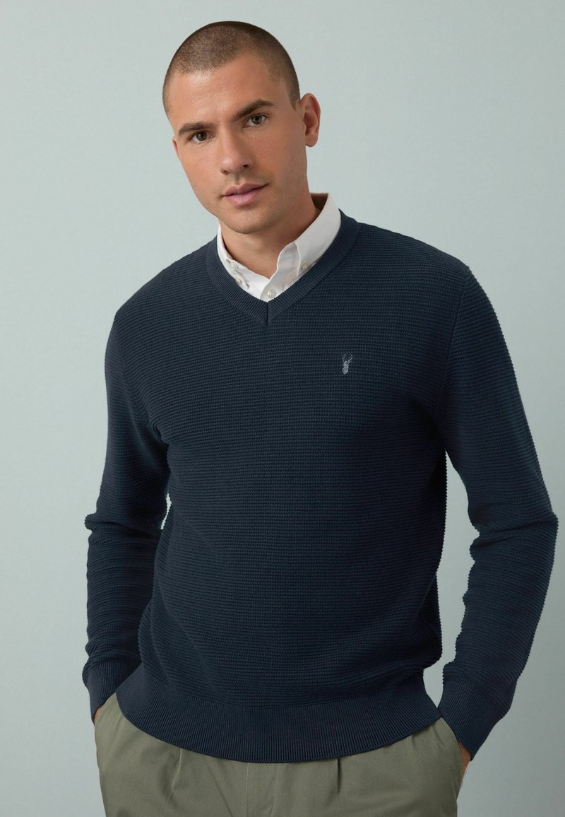 Marineblauwe gebreide trui met V-hals en lange mouwen; heeft een subtiele logo op de borst. Gedragen over een witte blouse.