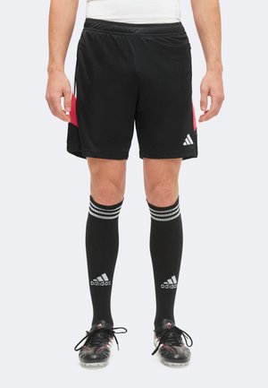 Osoba nosząca czarne sportowe spodenki Adidas, czarne skarpety Adidas do kolan z białymi paskami oraz czarne korki z czerwonymi detalami.