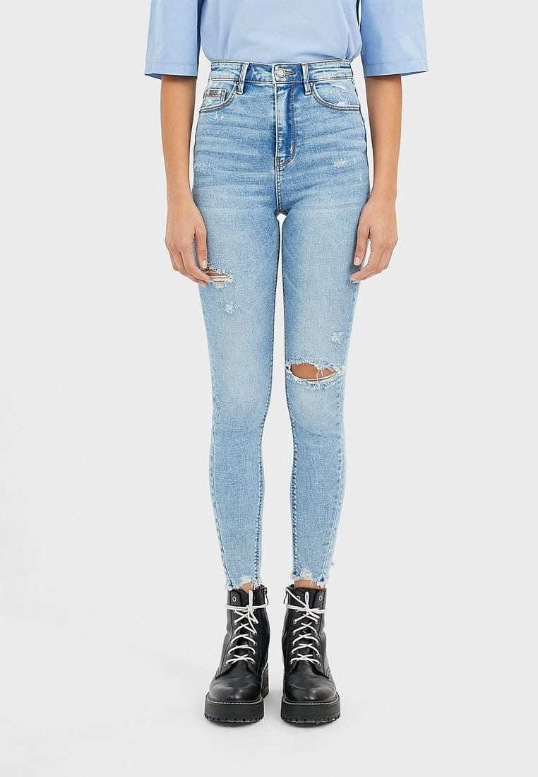 Jean skinny taille haute bleu clair avec déchirures usées et ourlets effilochés, associé à des bottines noires à lacets sur une personne.