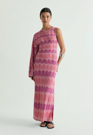 Mujer con un vestido largo sin mangas en tonos rosa y púrpura con patrón chevron, que tiene una manga larga y un puño plateado, de pie frente a un fondo liso.