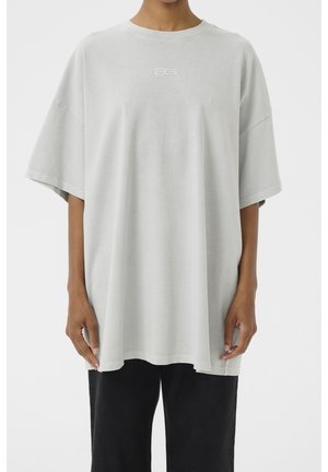 Person trägt ein übergroßes hellgraues T-Shirt mit kleinem weißen Logo auf der Brust und schwarze Hose, steht vor einem einfarbigen weißen Hintergrund.
