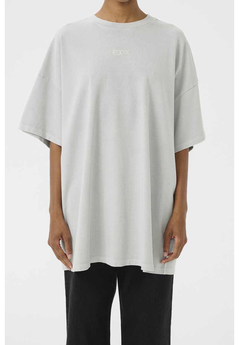 Personne portant un t-shirt gris clair oversize avec un petit logo blanc sur la poitrine et un pantalon noir, debout devant un fond blanc uni.