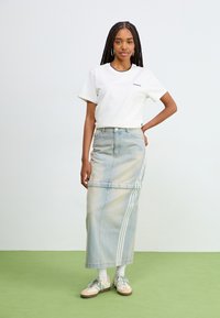 Hvit bomull t-skjorte og lys blå denim maxiskjørt med sidelister. Modellen har på seg pastellfargede joggesko og hvite sokker, og står på grønnt gulv.