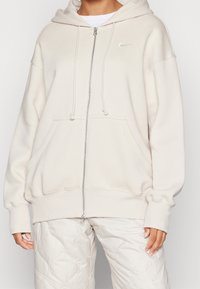 Krämfärgad zip-up hoodie i mjukt material med dragsko i huvan, kengurufickor och en liten silverlogo på bröstet.