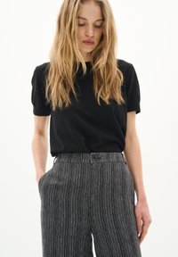 Schwarzes Kurzarmshirt, kombiniert mit hochgeschnittenen, gestreiften Schwarz-Weiß-Hosen mit Knopfverschluss und seitlichen Taschen.